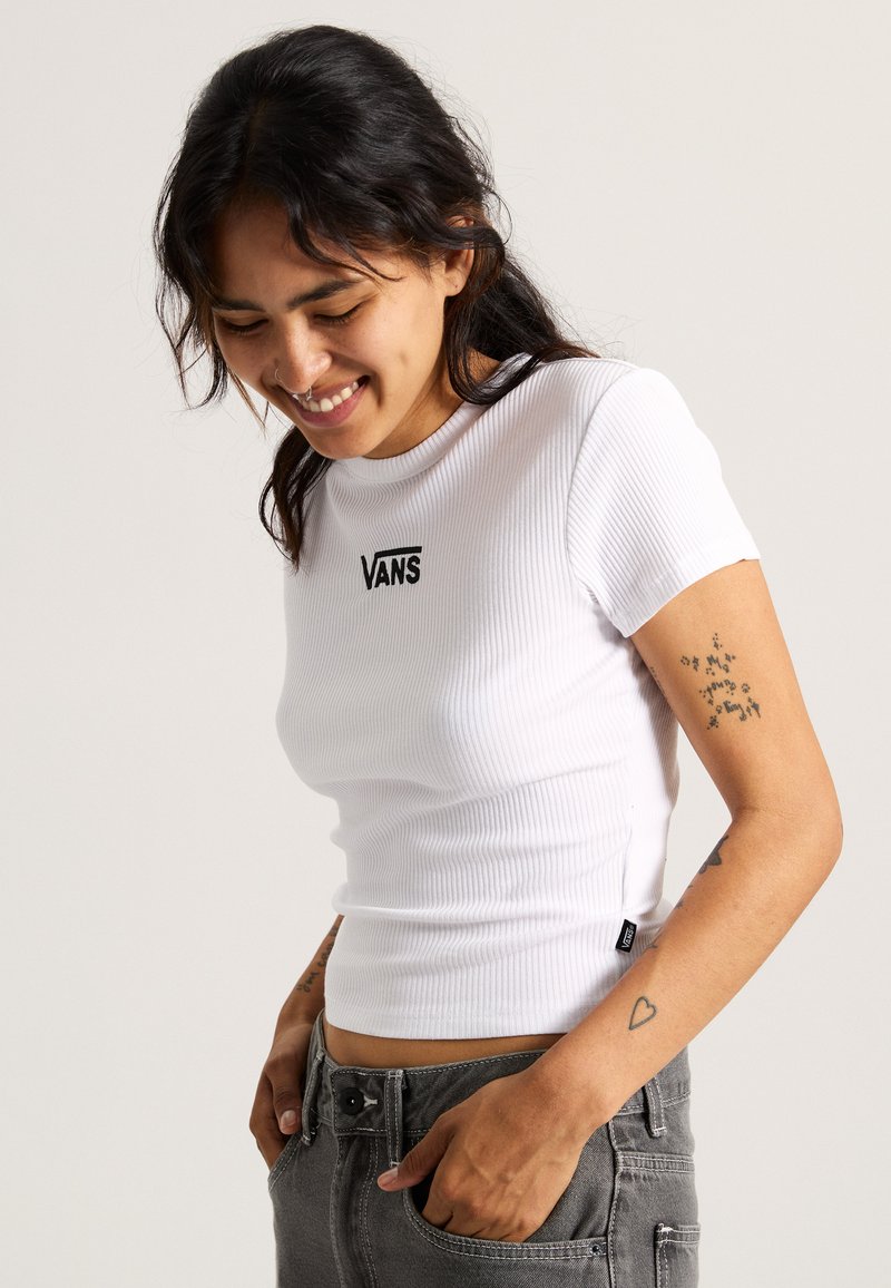 Hvid ribbet kortærmet t-shirt med Vans-logo, tætsiddende design, rund halsudskæring, parret med grå jeans, der har synlige sømme og lommer.