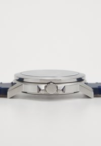 Montre en acier inoxydable argenté avec une lunette lisse, un cadran en verre rond et un bracelet en cuir navy texturé. Présente une couronne latérale avec un détail de logo.