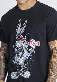Černé tričko s grafikou cartoon králíka s kostkovaným vzorem, bílými skvrnami a červeným štítkem "KAVAGH".