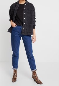 Schwarze Jeansjacke mit silbernen Knöpfen, getragen über einem schwarzen Hemd; kombiniert mit blauen, hochgekrempelten Jeans und braunen, schlangenmusterierten ankle Boots.