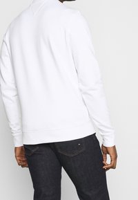 Felpa bianca con maniche lunghe, colletto rotondo e polsini a coste, indossata con jeans in denim scuro che presentano un piccolo logo sulla tasca posteriore.