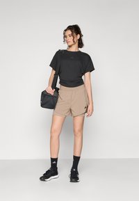 Maglietta atletica nera, pantaloni corti beige, calze nere e sneakers neri. Il modello tiene in mano una piccola borsa nera, con un logo discreto sui vestiti.