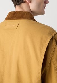 Giacca di cotone color giallo senape con colletto in velluto a coste. Presenta una finitura testurizzata e un'etichetta con logo cucita sul retro.