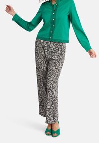 Chemise boutonnée verte avec des détails œillets, associée à un pantalon large à motifs floraux noir et blanc et des talons ouverts verts assortis.