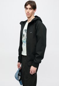 Veste à capuche noire avec fermeture éclair à l'avant, poignets élastiques et petit logo. Portée sur un t-shirt graphique vert clair et un pantalon rayé.