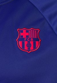 Emblema del FC Barcelona in rosso su un tessuto atletico blu, caratterizzato da un design a scudo con strisce e un motivo centrale di un pallone da calcio.