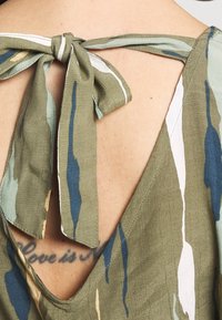 Green patterned fabric with a v-neckline and bow tie detail at the back; features abstract blue and beige stripes.
