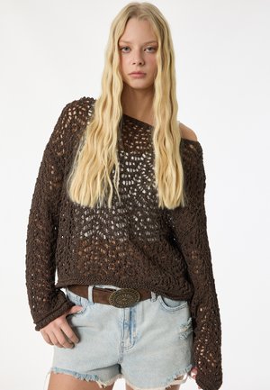 Giovane donna con lunghi capelli biondi che indossa un maglione largo marrone scuro a uncinetto e pantaloncini di jeans chiari con una cintura marrone.