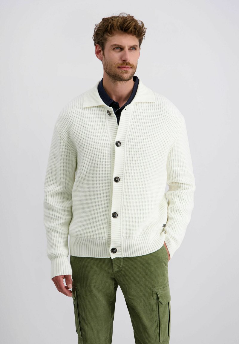 Weißer Strickcardigan mit einem strukturierten Muster, ausgestattet mit sechs dunklen Knöpfen und einem gerippten Kragen. Kombiniert mit grünen Cordhosen.