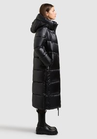 Lange zwarte pufferjas met een glanzende, gewatteerde textuur, voorzien van een capuchon, voorkruiszakken en een ritssluiting over de volle lengte. Gecombineerd met zwarte enkelboots.