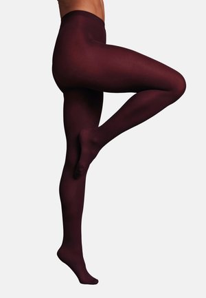 Personne portant des collants opaques bordeaux foncé, debout sur un pied avec l'autre jambe pliée et levée, sur un fond clair uni.