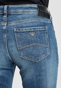 Modré džínové kalhoty mají zadní kapsu se zlatým emblémem, viditelné švy a texturovaný povrch. Poznámka o černé kožené cedulce na pásku.