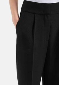 I pantaloni neri presentano una cintura ampia e piatta, un davanti plissettato e tasche laterali. Il materiale appare liscio con una leggera trama.