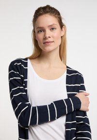 Cardigan navy con strisce bianche, in tessuto a maglia leggero, apertura frontale, maniche lunghe, indossato sopra una t-shirt bianca. Texture liscia, design casual.