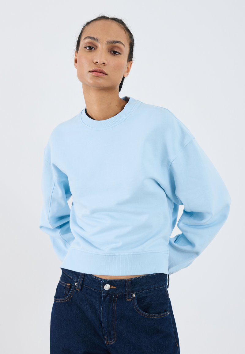 Sweatshirt cropped azul claro com gola redonda, punhos e bainha canelados. Combinado com jeans azul escuro com fecho de botão e bolsos.