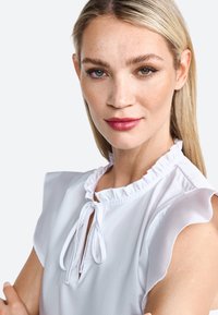 White blouse with ruffled neckline and tie detail. Short sleeves, lightweight fabric, smooth texture, and minimalistic design.