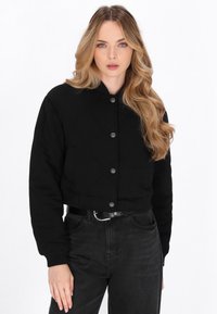 Schwarze, gekürzte, gesteppte Jacke mit rundem Kragen, Druckknöpfen und langen Ärmeln, kombiniert mit hoch geschnittenen schwarzen Denim-Jeans.