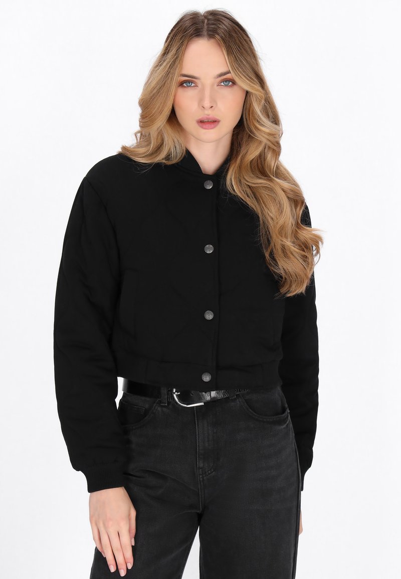 Schwarze, gekürzte, gesteppte Jacke mit rundem Kragen, Druckknöpfen und langen Ärmeln, kombiniert mit hoch geschnittenen schwarzen Denim-Jeans.