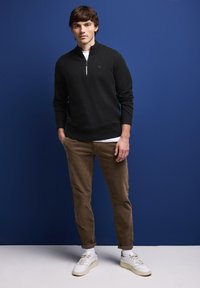 Zwart sweatshirt met ritssluiting, bruine corduroy broek en witte sneakers. Hete textuur, aansluitend ontwerp en zichtbaar logo op het sweatshirt.