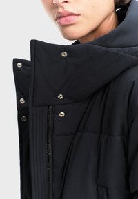 Čierna pufrová bunda s vysokým golierom, patentkami a zipsom vpredu. Mäkká, prešívaná textúra s hladkým povrchom.