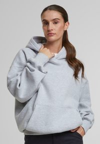 Šedá mikina s kapucňou a predným kengurím vreckom, s rebrovanými manžetami a vo voľnom strihu. Model drží kapucňu na neutrálnom pozadí.