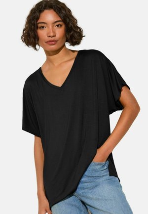 Lühikeste lokkis juustega naine, kandes lahtist musta V-kaelusega T-särki ja helesiniseid teksaseid, käsi taskus, tavalise taustaga.