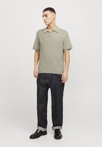 Polo de manga corta en color verde oliva con un tejido texturizado, con tapeta de tres botones, combinado con jeans de denim oscuro y zapatos negros.