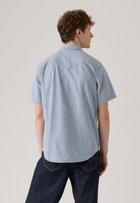 Πουκάμισο με κοντά μανίκια σε ανοιχτό μπλε χρώμα με λευκές κάθετες ρίγες, διαθέτει μυτερό γιακά, ραφή στον πίσω ώμο και στρογγυλεμένο τελείωμα.