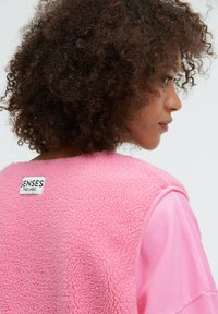 Rosa Fleece-Pullover mit strukturiertem Finish, unter dem sich ein glattes rosa Gewebe verbirgt, sowie einem schwarzen Etikett am Kragen.