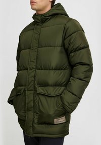 Polstret olivengrønn jakke med hette, to frontlommer og et lite merketlapp. Har en quiltet design med en glatt tekstur.