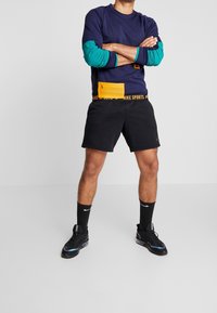 Maglietta a maniche lunghe navy con dettagli turchesi e tasca gialla. Pantaloni corti neri con marchio Nike. Sneakers nere con dettagli blu. Calze nere.
