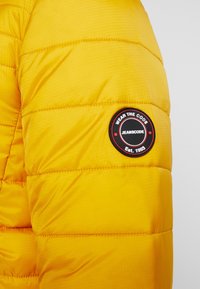 Gul pufferjakke-sektion med en quiltet tekstur. En rund sort plet viser "WEAR THE CODE JEANSCODE Est. 1993."