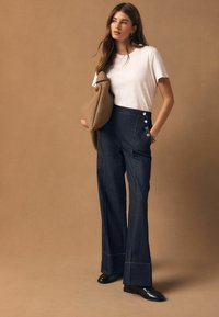 Donkerblauwe spijkerbroek met hoge taille, wijde pijpen, gouden stiksels en knoopdetails. Gecombineerd met een wit T-shirt en een beige trui met textuur.