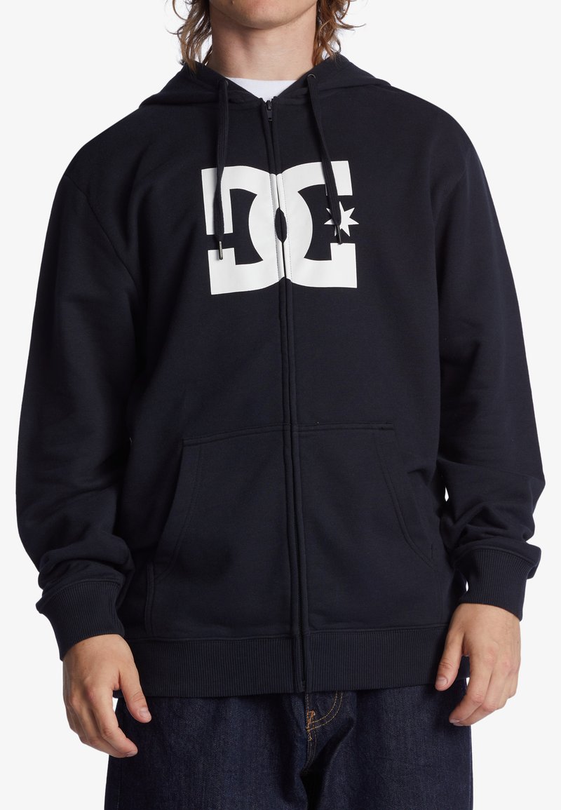 Navy blue zip-up hoodie made of soft material, featuring a large white DC logo on the chest, kangaroo pockets, and ribbed cuffs.