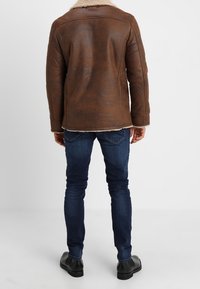 Brązowa skórzana kurtka z kołnierzem z owczej skóry, widocznymi szwami i prostym krojem, w zestawieniu z niebieskimi dżinsami i czarnymi butami.