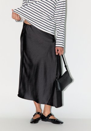 Femme portant une jupe midi en satin noir, un chemisier rayé à manches longues, des ballerines noires cloutées, et tenant un petit sac à main noir.