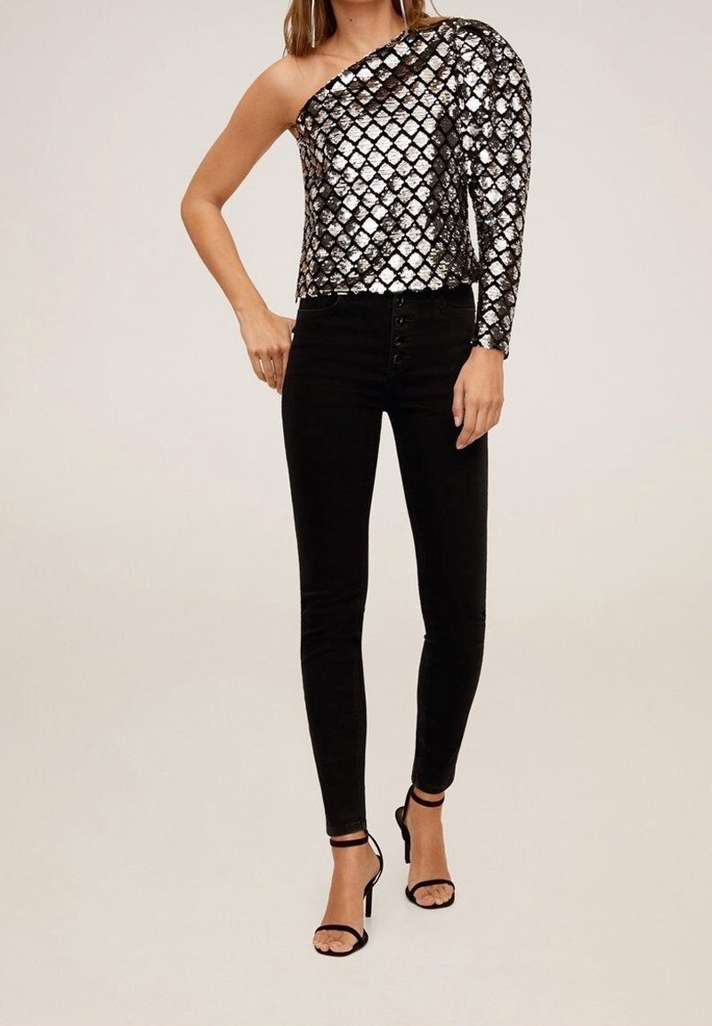 Femme portant un top argenté à paillettes une épaule, un pantalon skinny noir taille haute et des escarpins noirs à bride à la cheville, debout devant un fond uni.