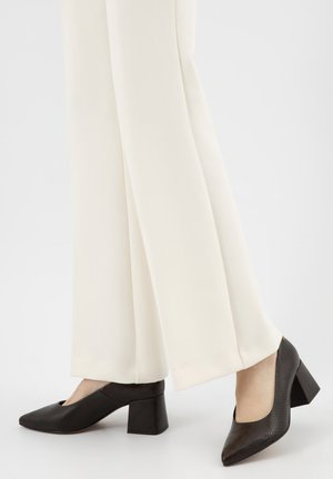 Frau trägt cremefarbene, weit geschnittene Hose und dunkelbraune, strukturierte Blockabsatzschuhe und steht auf weißem Boden.