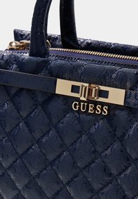 Granatowa torebka pikowana o teksturowanym wykończeniu, złotym okuciu oraz wyraźnym logo "GUESS" na ozdobnym pasku.