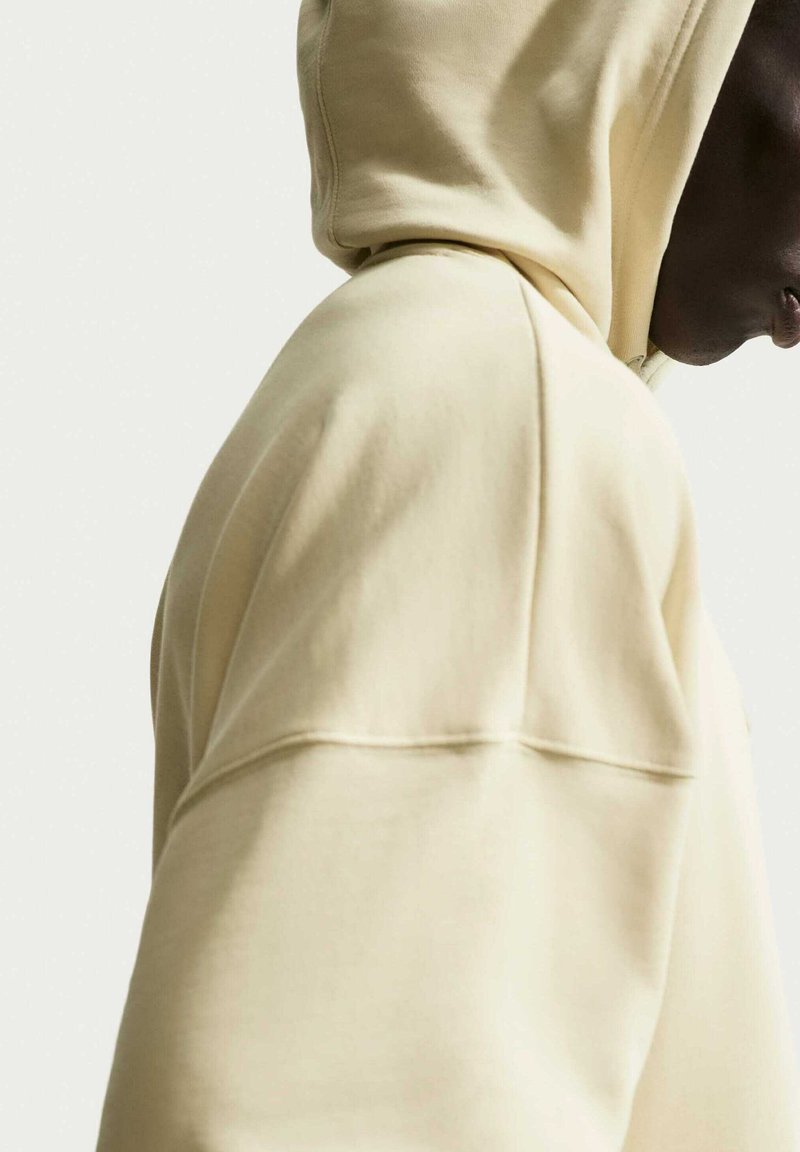 Personne en sweat à capuche crème vue de côté sur un fond uni clair, visage partiellement visible.