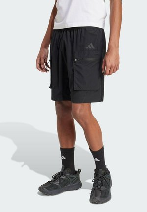 Mann trägt schwarze Sportshorts mit Reißverschlusstasche an der Seite, schwarze Adidas-Socken, schwarze Sneakers und ein weißes T-Shirt, steht vor hellem Hintergrund.