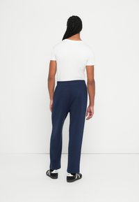 Braidelt hajjal rendelkező férfi fehér pólót, sötétkék nadrágot és fekete sportcipőt viselve áll egy sima fehér fal előtt.