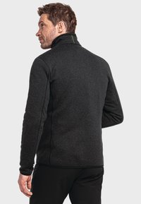 Schwarze Fleecejacke mit hohem Kragen, strukturiertem Oberflächen und kontrastierendem grünen Nähten auf dem Rücken, kombiniert mit schwarzen Hosen.