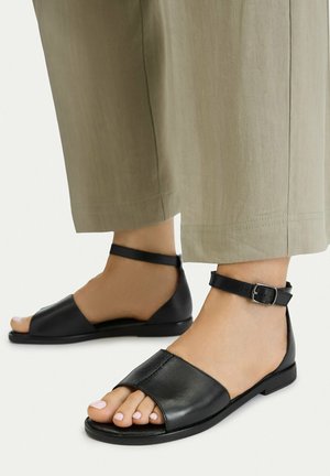 Nohy v černých otevřených sandálech s páskem kolem kotníku, doplněné béžovými širokými kalhotami s culíkem na světlém pozadí.