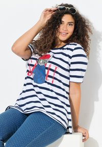 Gestreepte t-shirt in wit en marineblauw, met een kleurraf grafiek van een kat met een rode hoed. Draag het met blauwe broek met polka dots, relaxed fit.