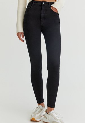 Vrouw draagt zwarte high-waist skinny jeans en witte sneakers, staand met één hand in de zak tegen een egale achtergrond.