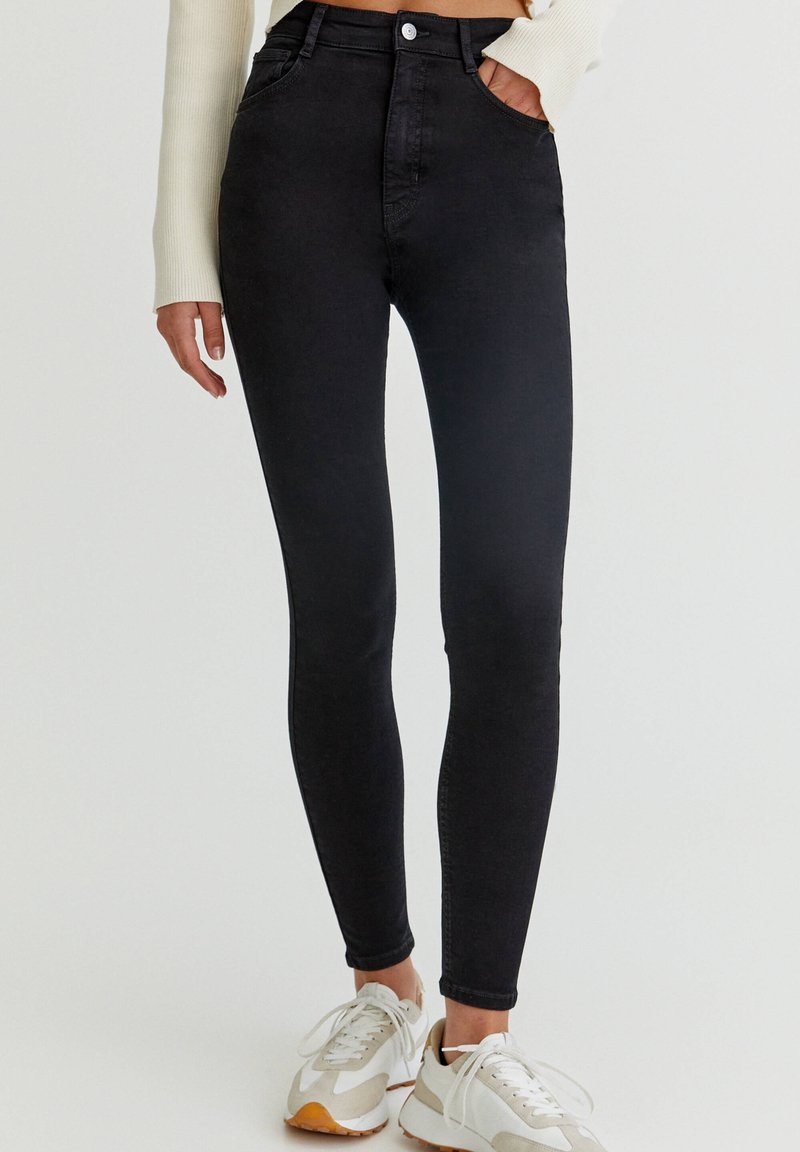 Femme portant un jean skinny noir taille haute et des baskets blanches, debout avec une main dans la poche devant un fond uni.