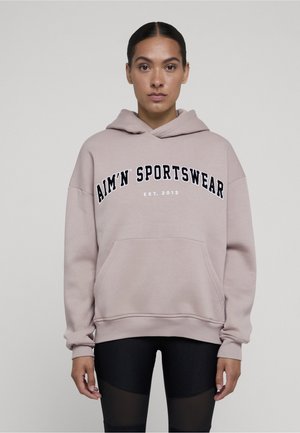 Person som har på seg en lysebeige hettegenser med teksten "AIM'N SPORTSWEAR EST. 2013" og svarte leggings med meshpaneler, står mot en ensfarget bakgrunn.