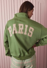 Woman with brown hair wearing a green sweatshirt with "PARIS" on the back and green-checkered pants, standing against a pink textured wall.