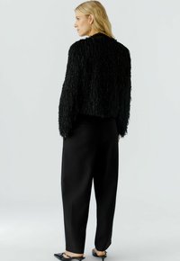 Frau, die nach links schaut, trägt einen strukturierten schwarzen Langarmtop, lockere schwarze Hosen und schwarze spitze Absatzschuhe mit einem Riemen.